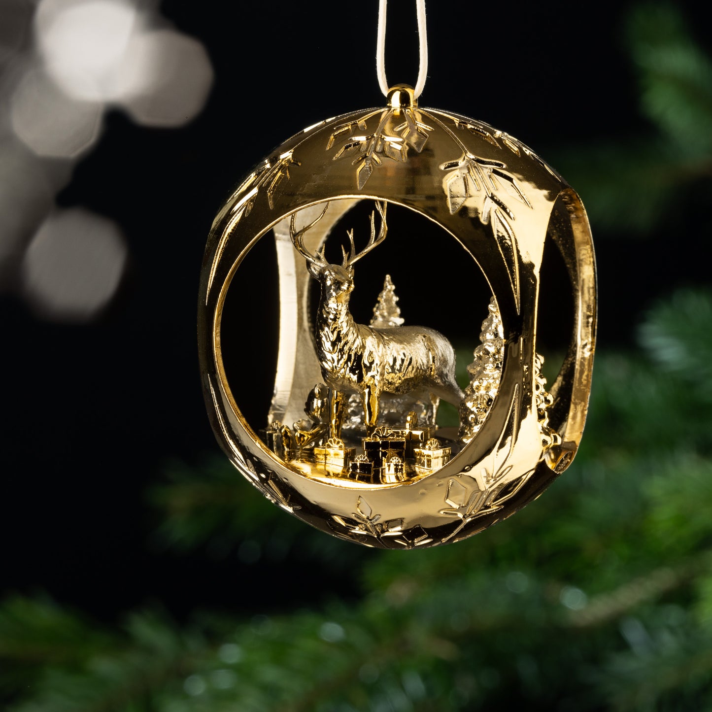 Auf dem Bild ist eine goldene Christbaumkugel zu sehen. Im Inneren der Kugel verzaubert dich ein filigraner Hirsch, der in einem verschneiten Wald steht. Geschenke um ihn herum und ein Teddybär, der im Schnee sitzt, sorgen für eine märchenhafte Atmosphäre.