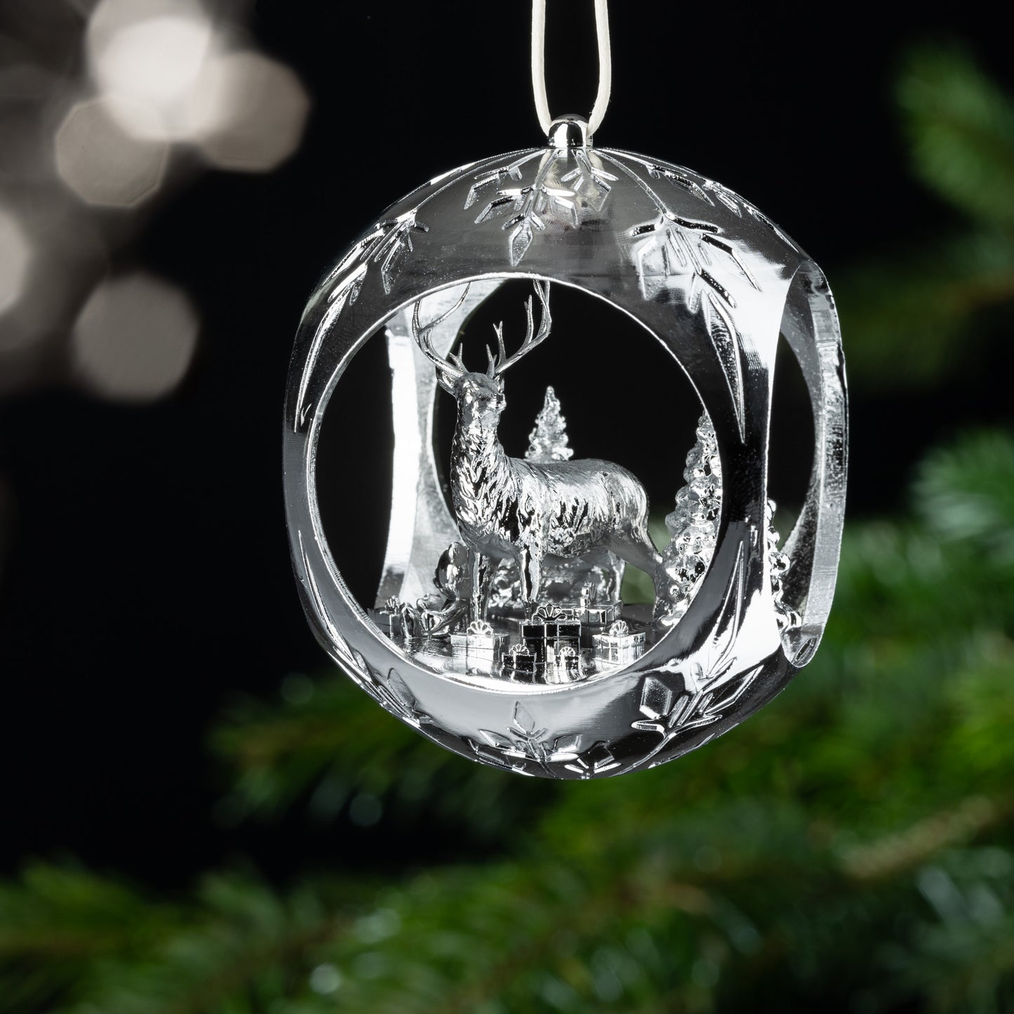 Auf dem Bild ist eine silberne Christbaumkugel zu sehen. Im Inneren der Kugel verzaubert dich ein filigraner Hirsch, der in einem verschneiten Wald steht. Geschenke um ihn herum und ein Teddybär, der im Schnee sitzt, sorgen für eine märchenhafte Atmosphäre.