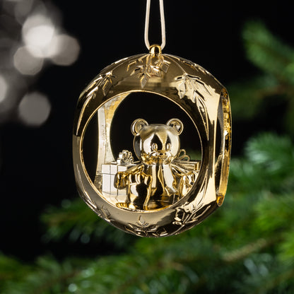 Auf dem Bild ist eine goldene Christbaumkugel zu sehen. Im Inneren der Kugel befindet sich ein großer Teddybär, der gemütlich im Schnee sitzt, umgeben von Geschenken. Diese Szene strahlt Gemütlichkeit und Freude aus.