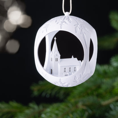 Auf dem Bild ist eine weiße Christbaumkugel zu sehen. Im Inneren der Kugel erhebt sich eine idyllische Kirche mit einem Kreuz, umgeben von Bäumen und einladenden Bänken. Diese friedliche Szenerie strahlt Ruhe und Geborgenheit aus.